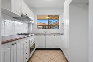 Kitchen with white cabinetry, tiled splashback, gas cooktop, oven, and window overlooking neighbouring rooftops at 13/14–16 Cairns Street, Riverwood NSW.