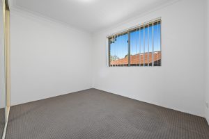 Bedroom with grey carpet, white walls and window with vertical blinds at 13/14–16 Cairns Street, Riverwood NSW 2210.