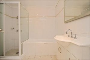 Clean apartment bathroom with white tiled walls, glass shower enclosure, built-in bathtub, and vanity unit at 8/13–19 Hogben Street, Kogarah NSW 2217