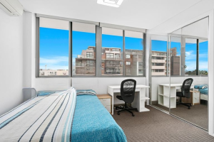 Bright bedroom with single bed, study desk, mirrored wardrobe, and large windows overlooking surrounding buildings at 712/12 Bonar Street, Arncliffe NSW 2205