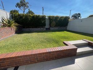Level, grassed backyard with brick retaining walls and paved steps at 31A Lawrence St, Peakhurst NSW 2210