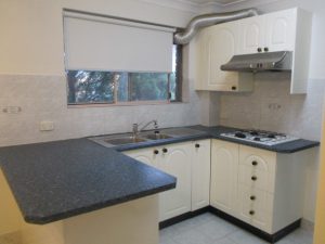 Kitchen at 10/50 Melvin Street, Beverly Hills NSW 2209 showing an L-shaped benchtop, stainless steel sink, gas cooktop, rangehood, white cabinetry, and window with roller blind